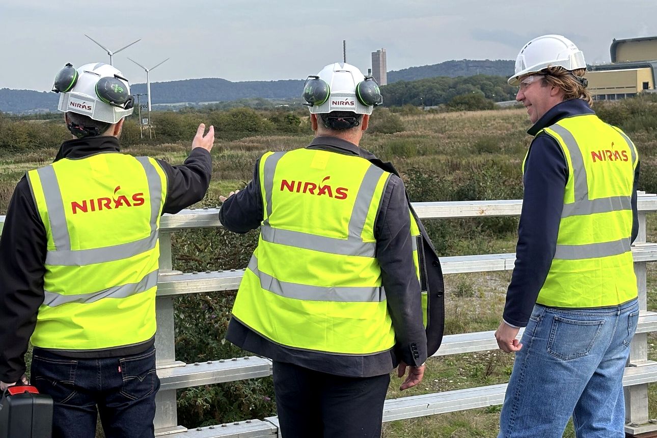 Three members of the site visit team in NIRAS high-visibility vests and hard hats, one pointing across the site, with wind turbines and industrial buildings in the background