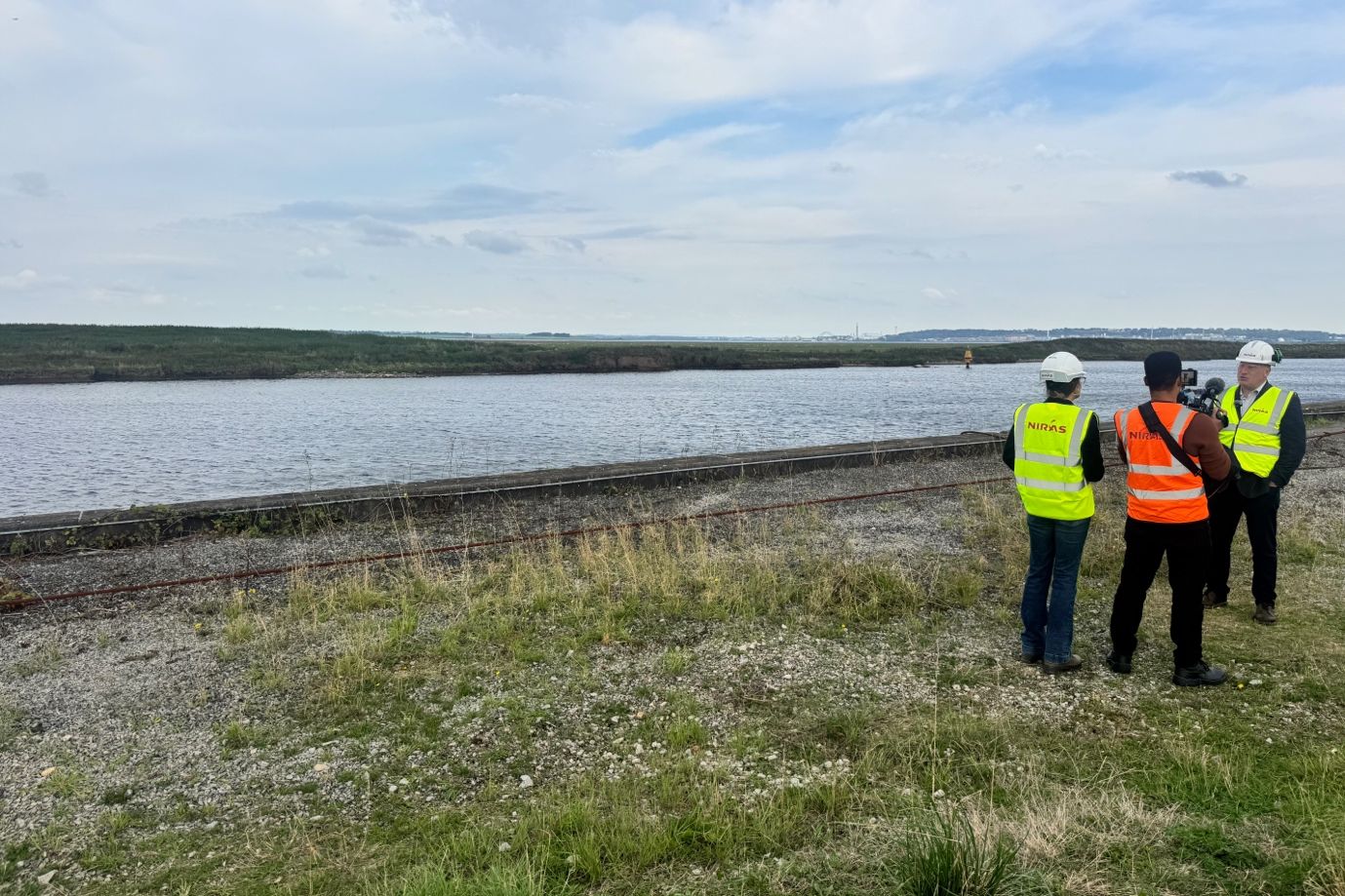 Three people in high-visibility vests and hard hats beside a wide stretch of water, one filming with a video camera