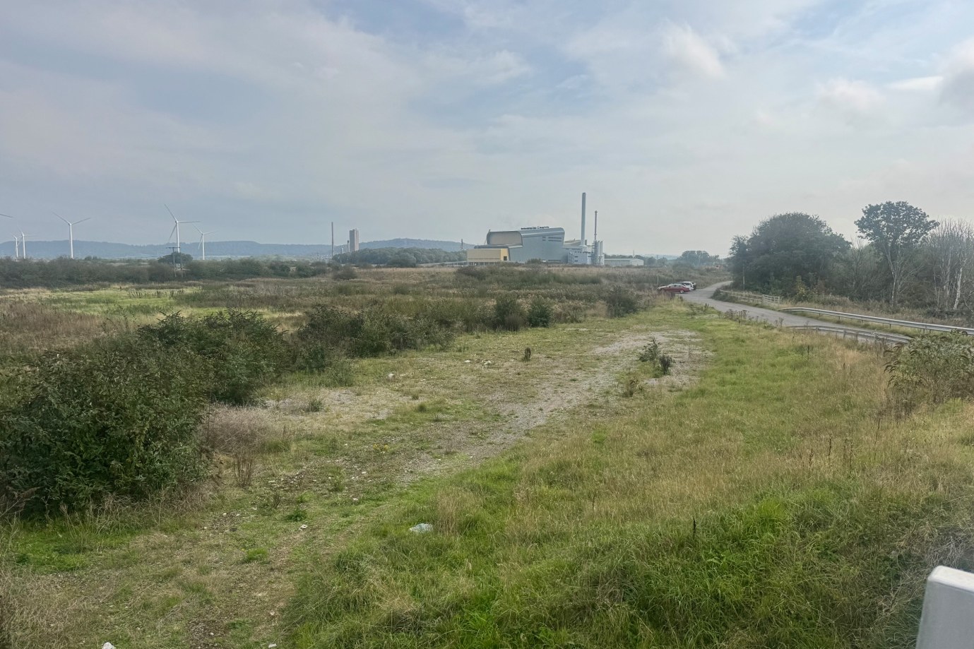 Open, overgrown land beside a road, with industrial buildings, a chimney stack and wind turbines on the horizon